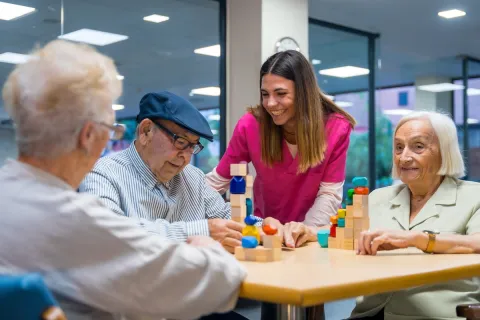 jeune soignante souriante aide trois personnes âgées à empiler blocs colorés.